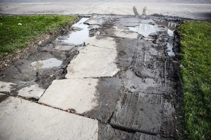 Broken sidewalk with large cracks and puddles beside a grassy verge
