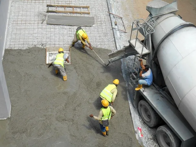 Workers in yellow vests pour concrete beside a cement truck at a construction site.