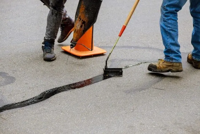 Workers repairing a cracked asphalt road beside orange cones, with a shovel and brooms visible